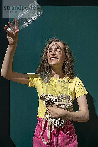 Lächelnde Frau  die an einem sonnigen Tag die Augen abschirmt und einen Netzbeutel und eine Plastikflasche in der Hand hält