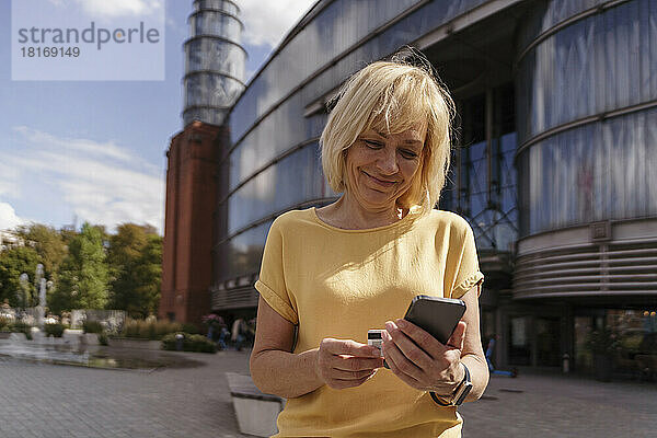 Lächelnde Frau mit Kreditkarte beim Online-Einkauf per Smartphone an einem sonnigen Tag