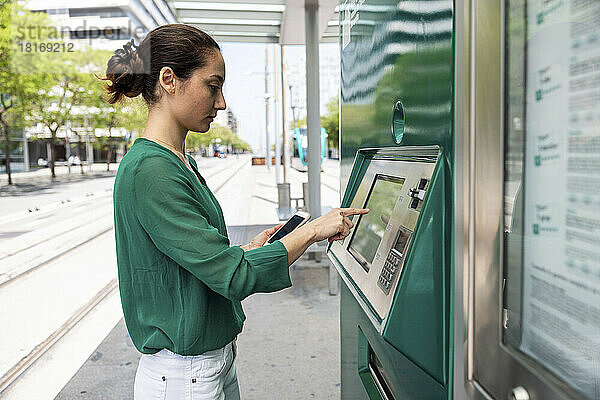 Frau mit Smartphone benutzt Fahrkartenautomat an Straßenbahnhaltestelle