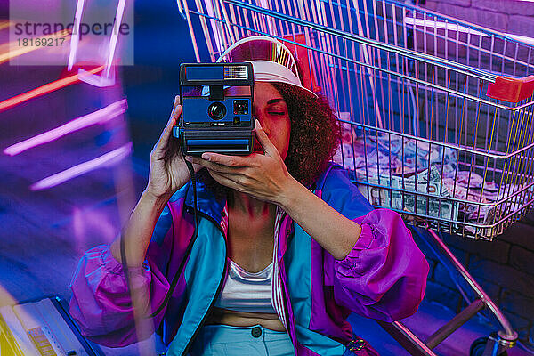Junge Frau fotografiert mit Sofortbildkamera vor dem Einkaufswagen