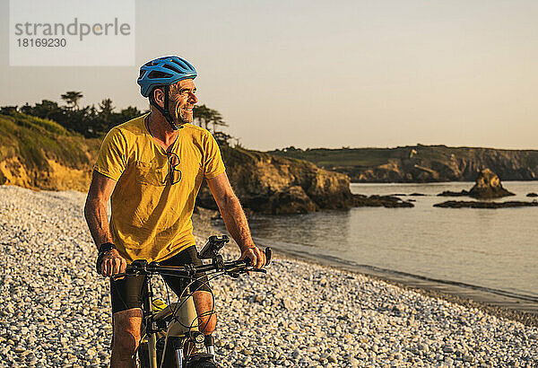 Lächelnder reifer Mann  der bei Sonnenuntergang am Ufer Fahrrad fährt