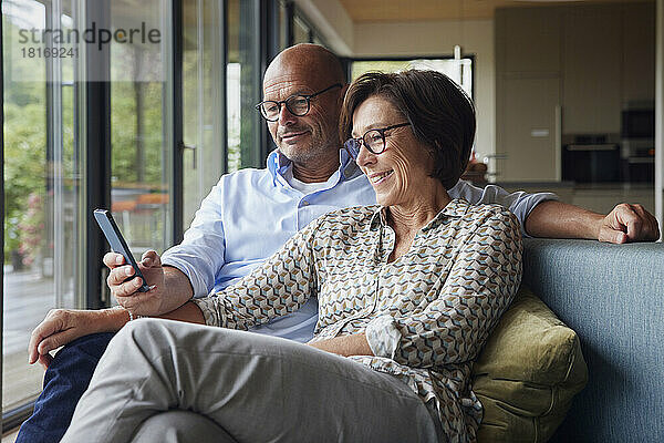 Glückliche Frau mit Mann  der zu Hause auf dem Sofa ein Smartphone benutzt