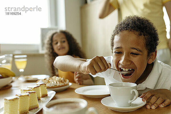 Glücklicher Junge trinkt heiße Schokolade mit Löffel von Schwester am Esstisch