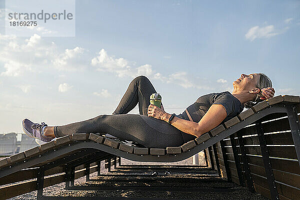 Frau hält Wasserflasche in der Hand und entspannt sich an einem sonnigen Tag auf einer Holzbank