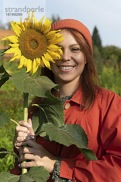 Lächelnde Frau mit braunen Haaren hält Sonnenblume im Garten