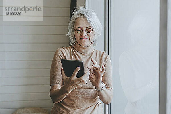Lächelnde reife Frau  die am Fenster steht und einen Tablet-PC benutzt