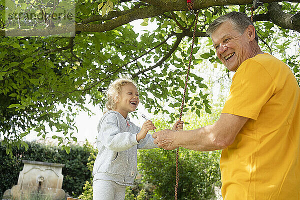Glücklicher älterer Mann mit Enkelin beim Schaukeln mit Seil im Garten