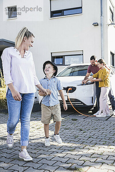 Glückliche Mutter blickt ihren Sohn an  während Vater und Tochter ein Elektroauto aufladen