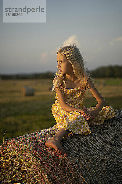 Girl with blond hair sitting on haystack at sunset