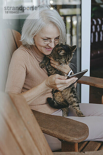 Happy mature woman sitting with cat and using tablet PC at home