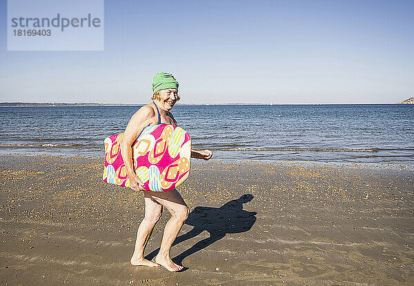 Glückliche ältere Frau läuft mit Surfbrett am Strand