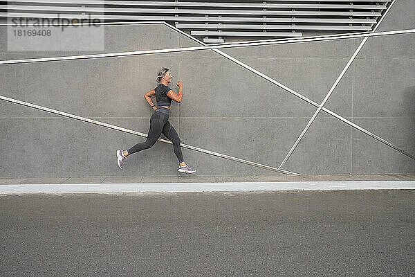 Reife Frau joggt auf Fußweg an grauer Wand