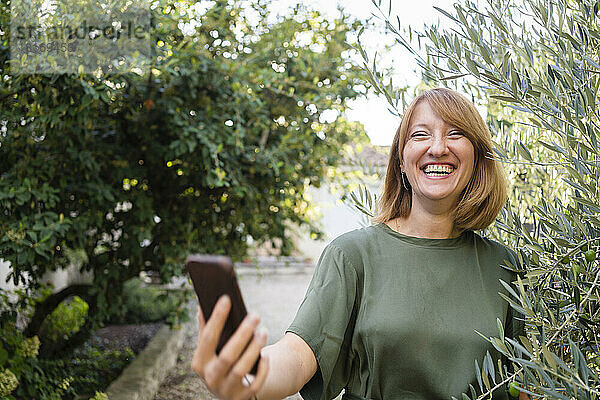 Fröhliche Frau mit Handy macht Selfie im Garten