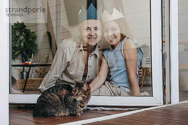 Glückliche Großmutter und Enkelin mit Krone sitzen zu Hause gemeinsam hinter einem Glasfenster