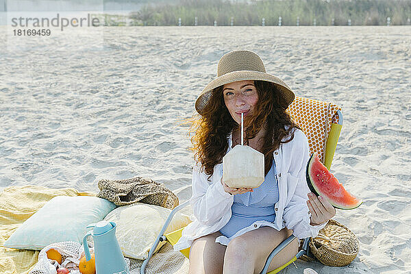 Frau mit Hut trinkt Kokosnusswasser am Strand