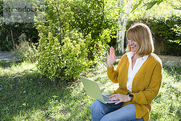 Lächelnder Freiberufler winkt durch seinen Laptop und sitzt im Garten