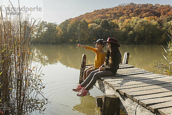 Junge zeigt auf Mädchen  das zusammen auf Steg am See sitzt