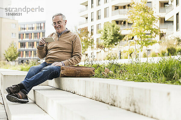 Glücklicher älterer Mann sitzt auf der Treppe und nutzt sein Smartphone