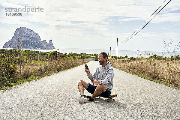 Glücklicher Mann mit Smartphone auf Skateboard sitzend