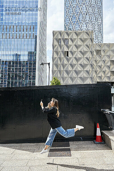 Glückliche Frau mit Smartphone springt an einem sonnigen Tag vor Gebäuden