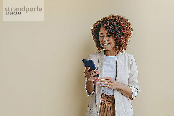 Lächelnde junge Geschäftsfrau  die ihr Mobiltelefon vor beigem Hintergrund benutzt