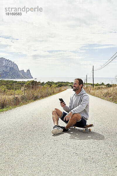 Glücklicher Mann mit Smartphone sitzt auf Skateboard