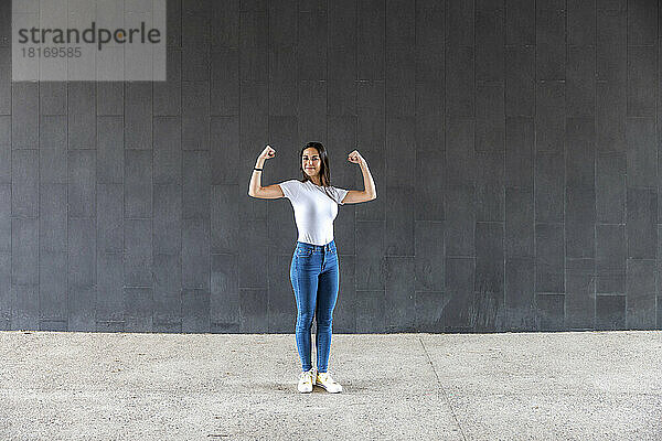 Lächelnde Frau  die auf dem Fußweg vor einer grauen Wand ihre Muskeln spielen lässt