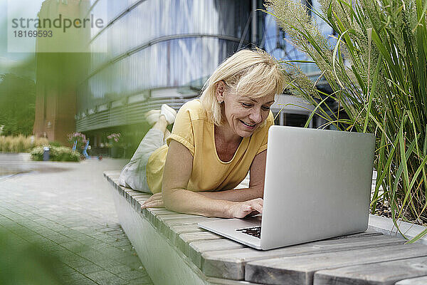 Happy businesswoman using laptop lying on bench