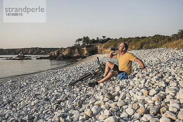 Mann trinkt Wasser mit dem Fahrrad und sitzt auf Kieselsteinen am Strand