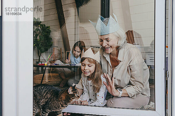 Großmutter und Enkelin tragen eine Krone und schauen durch ein Glasfenster auf die Katze