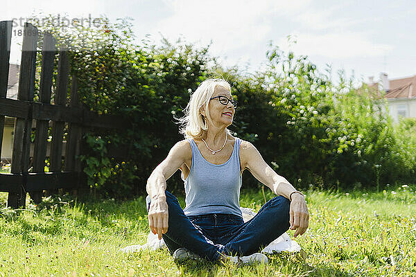 Lächelnde Frau sitzt an einem sonnigen Tag mit gekreuzten Beinen im Garten