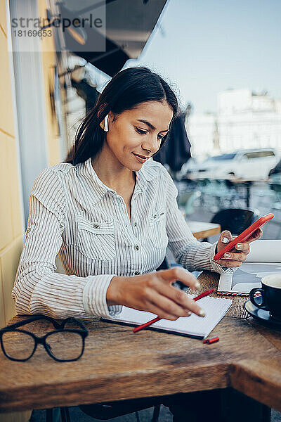 Lächelnde Geschäftsfrau mit Notizblock und Smartphone sitzt im Straßencafé