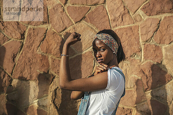 Junge Frau  die an einem sonnigen Tag an der Wand steht und ihre Muskeln spielen lässt