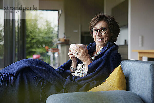 Glückliche Frau mit Kaffeetasse und Decke sitzt zu Hause auf dem Sofa