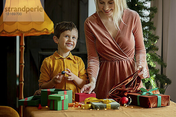 Glücklicher Junge mit Mutter  die zu Hause Weihnachtsgeschenke auf dem Tisch verpackt