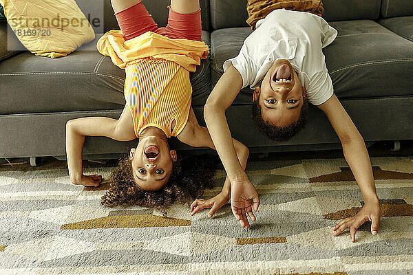 Girl and boy sticking out tongue enjoying upside down on sofa at home