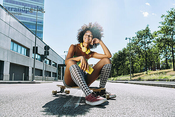 Lächelnde junge Frau mit Smartphone sitzt auf Skateboard