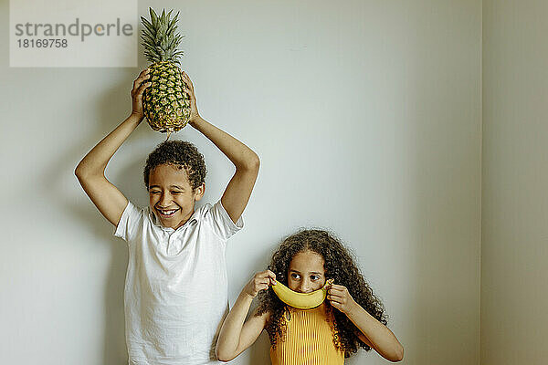Happy boy holding pineapple above head by sister covering lips with banana in front of wall