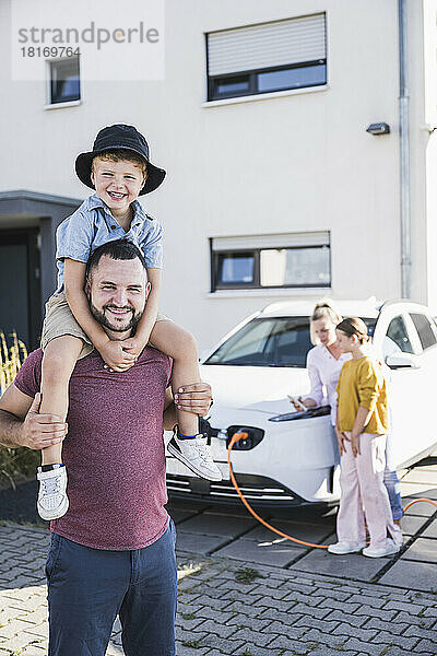 Glücklicher Vater trägt Sohn auf Schultern  während Mutter und Tochter Elektroauto aufladen