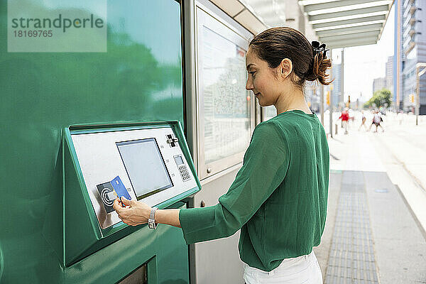 Lächelnde Frau  die an der Straßenbahnhaltestelle einen Fahrkartenautomaten mit Kreditkarte bedient
