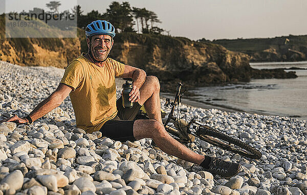 Glücklicher Mann sitzt mit dem Fahrrad und hält eine Wasserflasche am Strand