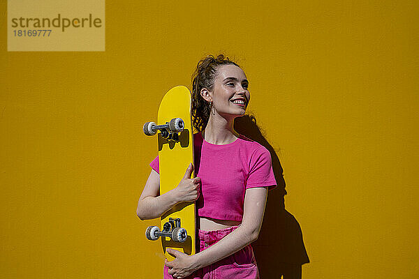 Nachdenkliche Frau hält an einem sonnigen Tag Skateboard vor der Wand