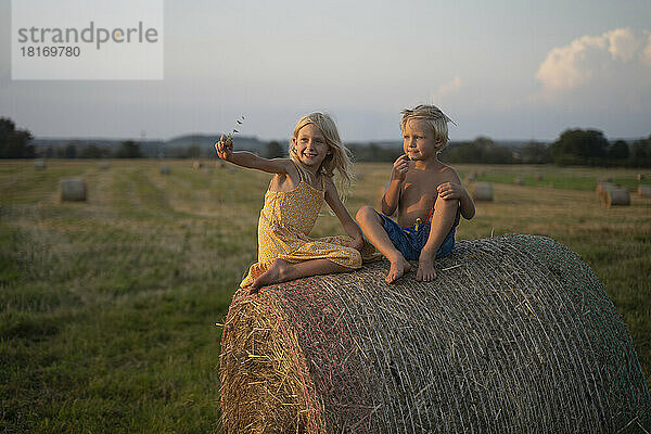 Smiling girl with arm outstretched sitting by brother on haystack