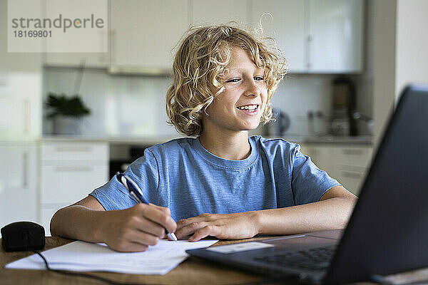 Fröhlicher Junge macht Hausaufgaben und schaut zu Hause auf den Laptop