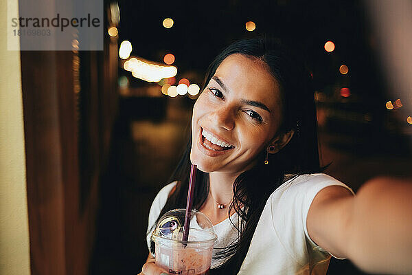 Cheerful woman with smoothie taking selfie at night