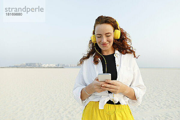Lächelnde Frau mit Kopfhörern und Smartphone am Strand