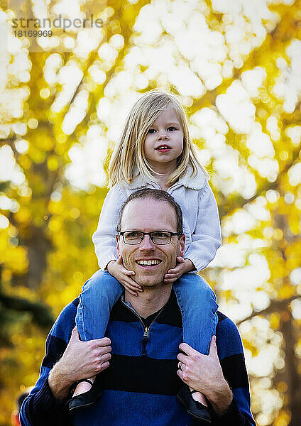 Ein Vater trägt seine kleine Tochter auf den Schultern  während er im Herbst in einem Stadtpark spazieren geht; St. Albert  Alberta  Kanada