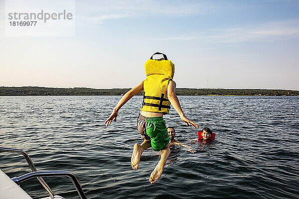 Junge  der von einem Boot springt und in einem See schwimmt  mit einer Schwimmweste  während sein Vater und sein Bruder ihn beobachten  Lac Ste. Anne; Alberta Beach  Alberta  Kanada