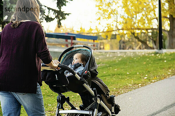 Eine Mutter geht mit ihrem Baby mit Down-Syndrom in einem Kinderwagen entlang eines Flusses in einem Stadtpark mit einer Brücke im Hintergrund während der Herbstsaison; St  Albert  Alberta  Kanada