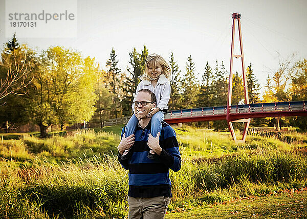 Ein Vater trägt seine kleine Tochter auf den Schultern  während er im Herbst in einem Stadtpark spazieren geht; St. Albert  Alberta  Kanada